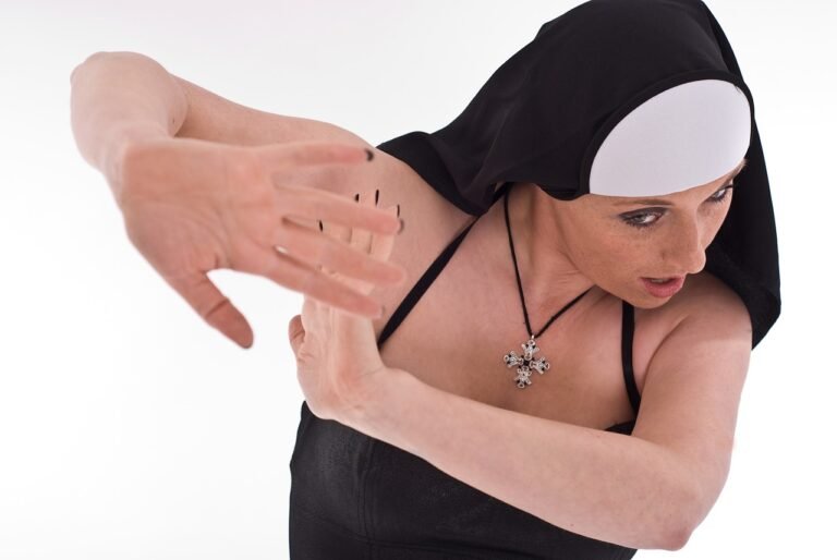 A woman wearing a black nun costume with a white headpiece, performing a dance pose with her hands raised.
