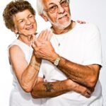 An elderly couple smiling and posing closely together against a plain background.