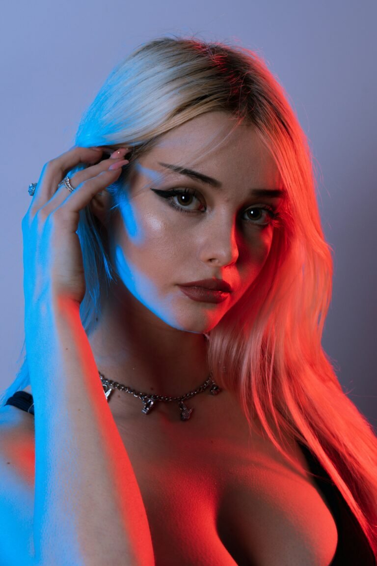 Portrait of a woman with long blonde hair under blue and red lighting, wearing a necklace and posing with one hand on her face.